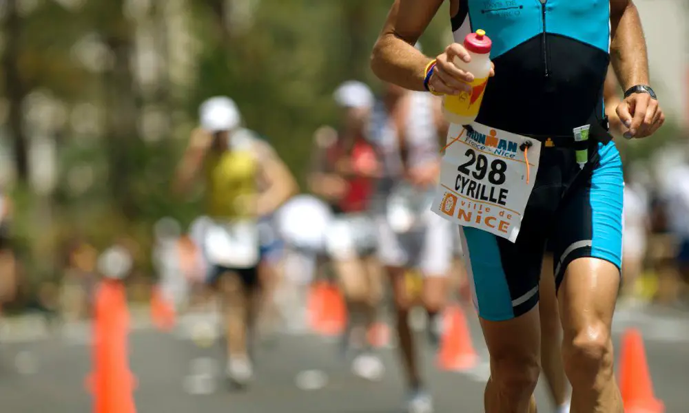 Marathon runner wearing blue one piece bathing suit holding a bottle of water with three runners behind him on the street