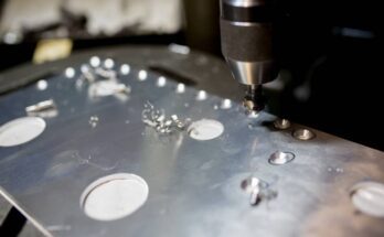 A machine drilling circular holes into a sheet of metal. The resulting metal burrs are off to the side.