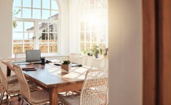 A sunroom with white walls, arched windows, and a wood dining room table with eight woven dining room chairs.