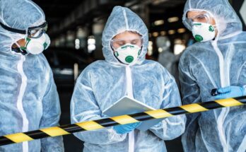 Three workers wearing HAZMAT suits are looking at information on a digital tablet and standing behind a pole barricade.