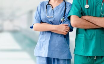 Two medical professionals wearing scrubs stand side-by-side with arms crossed in a hospital hallway.