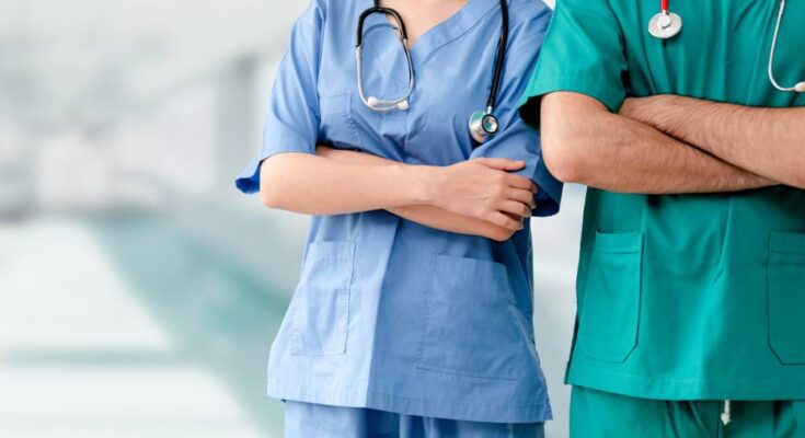 Two medical professionals wearing scrubs stand side-by-side with arms crossed in a hospital hallway.