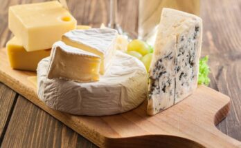 Six different types of hard and soft cheeses all organized on a wooden cutting board and ready to be paired with other foods.