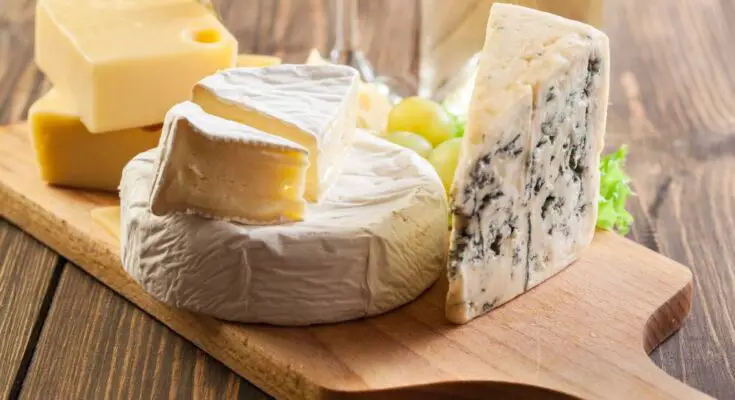 Six different types of hard and soft cheeses all organized on a wooden cutting board and ready to be paired with other foods.
