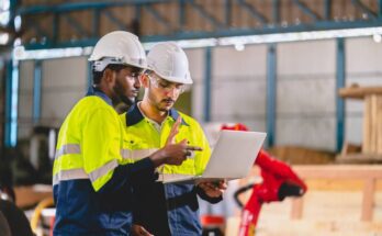 Two men standing in white hard hats and high-vis clothing working with a laptop on a manufacturing floor.