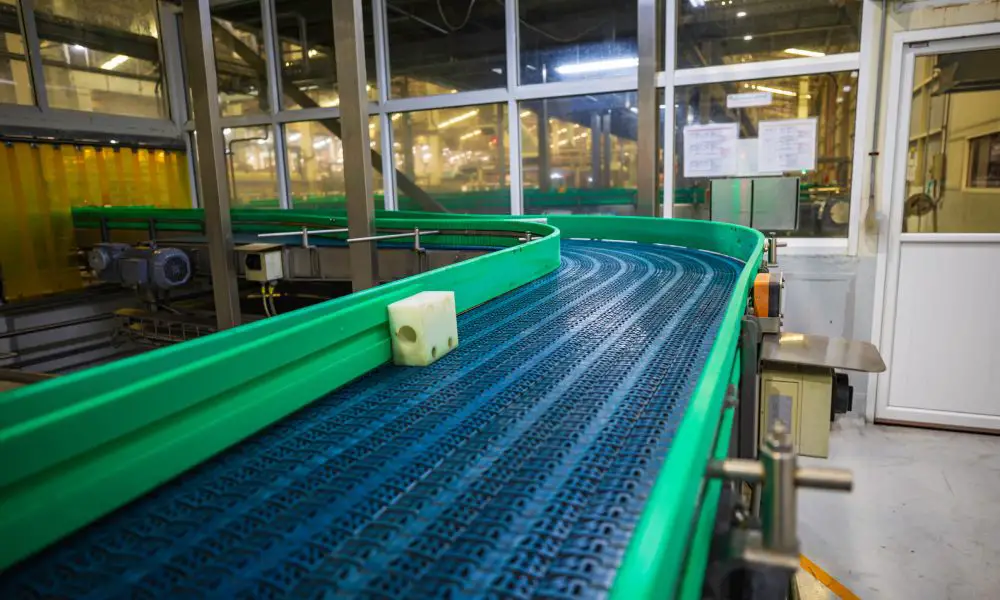 A black plastic conveyor belt with green plastic bumpers winding around a corner in a large factory.