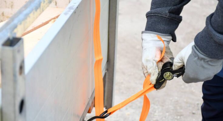 A logistics professional wearing work gloves holds a tensioning device to secure orange rachet straps to a truck trailer.