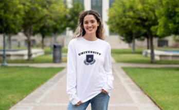 A first-year student standing in front of a campus building while wearing a long-sleeved university shirt.