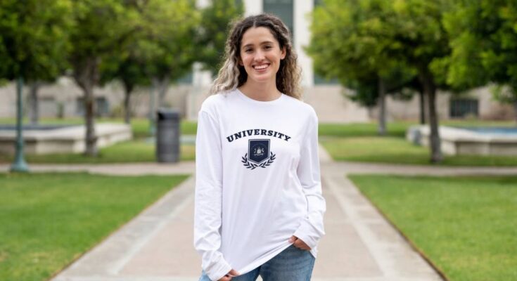 A first-year student standing in front of a campus building while wearing a long-sleeved university shirt.