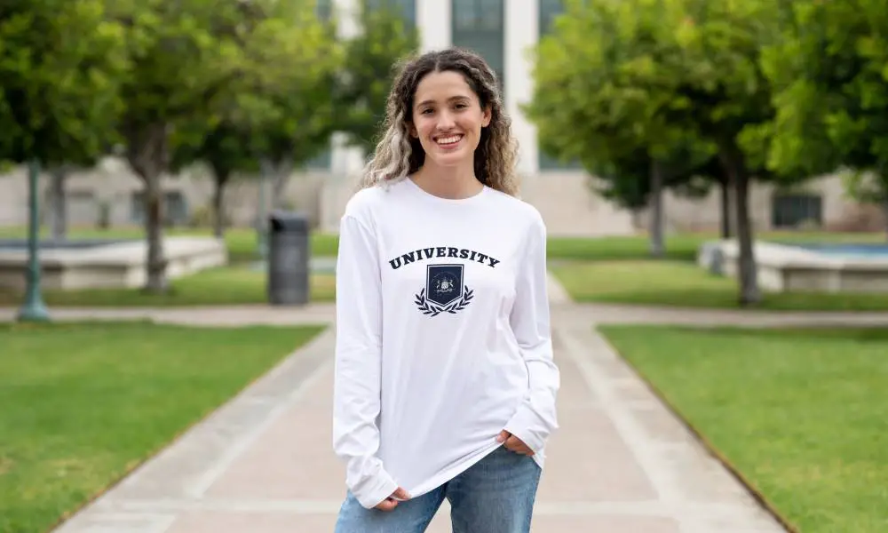 A first-year student standing in front of a campus building while wearing a long-sleeved university shirt.