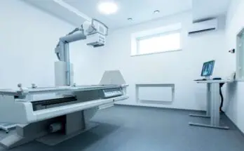 An immaculate and brightly lit radiology room in a hospital featuring an X-ray table and machine and computer monitor.