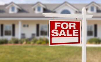 A red sign that reads "For Sale" posted in a yard with a long, one-story home blurred in the background.