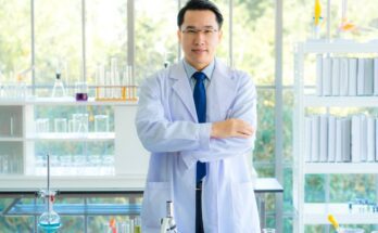 A lab manager stands in his laboratory with his arms folded. He wears a white coat, a blue tie, and glasses.