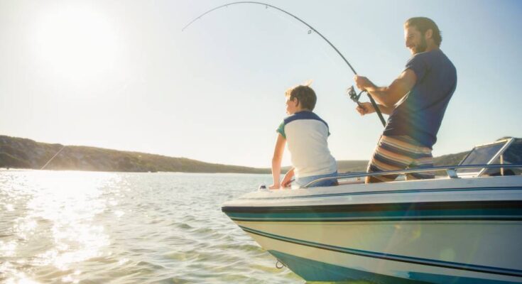 A father and son standing on a boat that is floating in a lake. The father is reeling in a catch with his fishing pole.