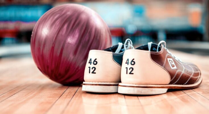 A red bowling ball and a pair of size 12 bowling shoes sitting on the ground in front of a bowing lane.