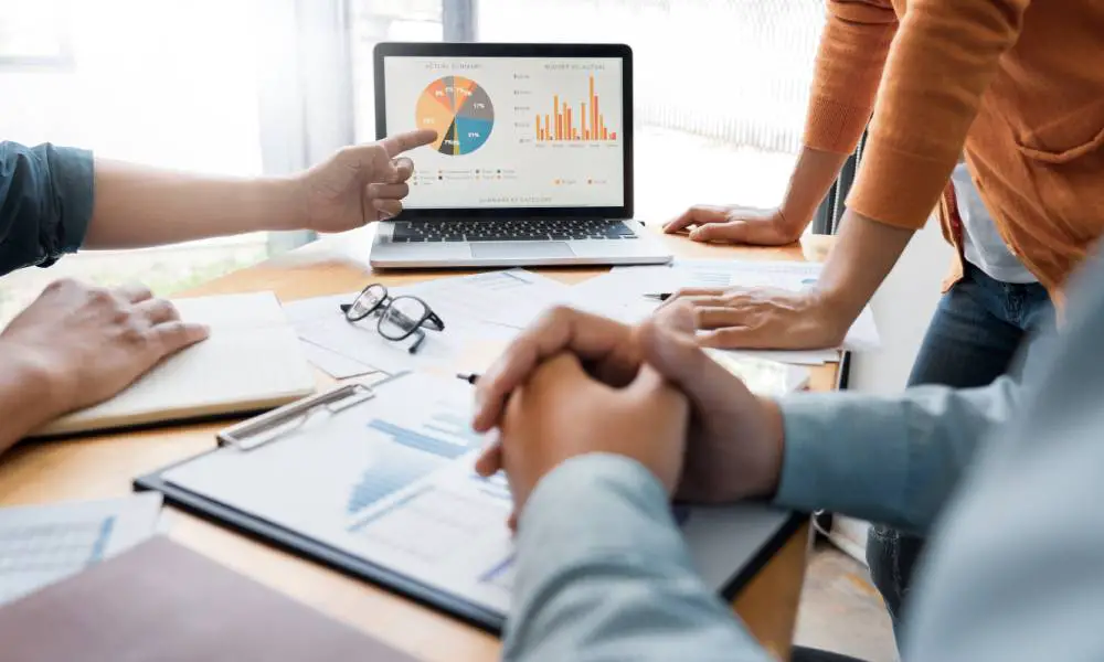 A diverse group of coworkers gathered around a table and focused on a laptop screen displaying charts and graphs.