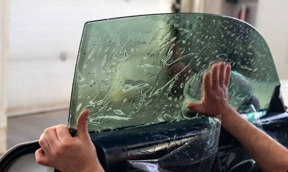 A person is applying a sheet of tint to a vehicle's driver's side window. A hand is smoothing the bubbles.