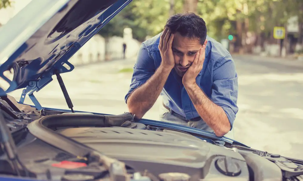 A man in a blue shirt holding his head in his hands and making a worried expression while looking at his car's engine.