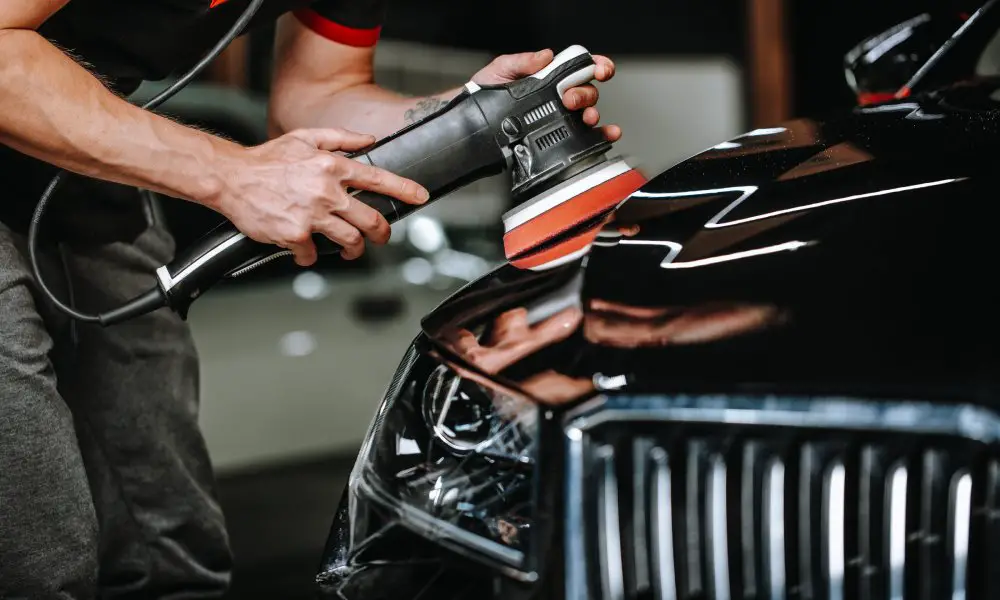 A man holding an orbital polisher against a sleek black car in a car studio. He is detailing and cleaning the car.