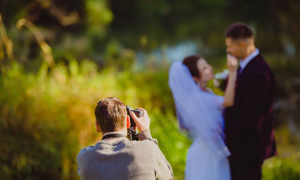 A photographer snaps pictures of a bridal couple outside in a green park. Only the photographer is in focus.