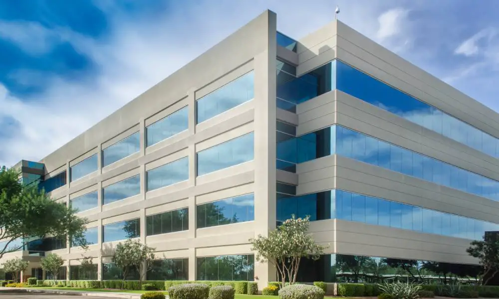 A gray, four-story commercial building stands high, surrounded by a blue sky. It has a parking lot, trees, and shrubs.