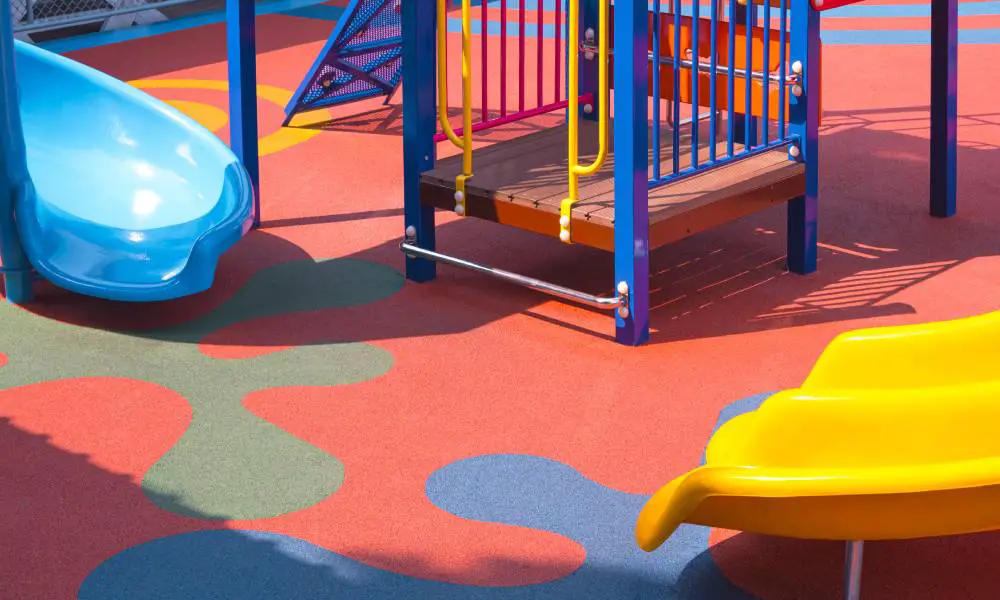 A playground with a blue slide and a yellow side, climbing equipment, and a rubber surface. The surface is red with blue designs.
