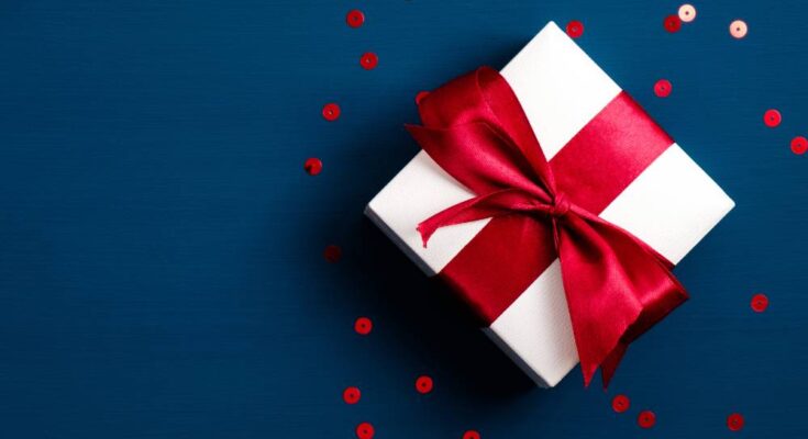 A white gift box tied with a red ribbon and red circular confetti on a dark blue background.