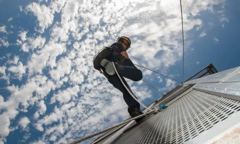 A firefighter using rope to rappel down the side of a fire station tower. The sky is blue and cloudy in the background.
