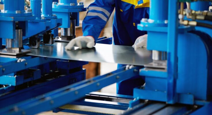 A worker dressed in a blue and yellow uniform is holding a piece of sheet metal in between a large blue machine.
