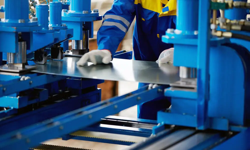 A worker dressed in a blue and yellow uniform is holding a piece of sheet metal in between a large blue machine.