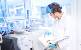 A scientist stands in a lab and holds a manual for a piece of equipment. He wears a shield over his face and blue gloves.