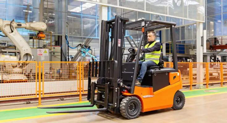 A man wearing a reflective safety vest is driving an orange and black forklift through a large warehouse.