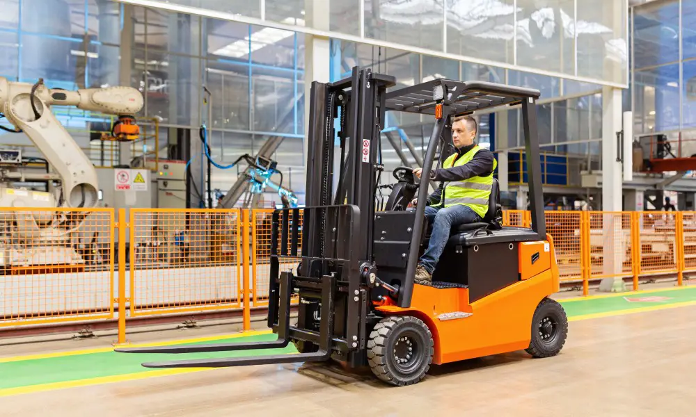 A man wearing a reflective safety vest is driving an orange and black forklift through a large warehouse.