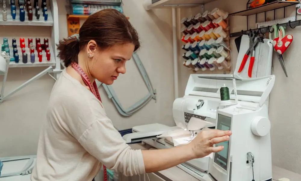 A woman in a white shirt with a pink tape measure around her neck. She's working on the screen of her embroidery machine.
