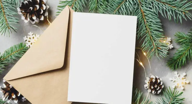 A close-up of a blank holiday card and envelope surrounded by pine tree branches, pine cones, and snowflake lights.