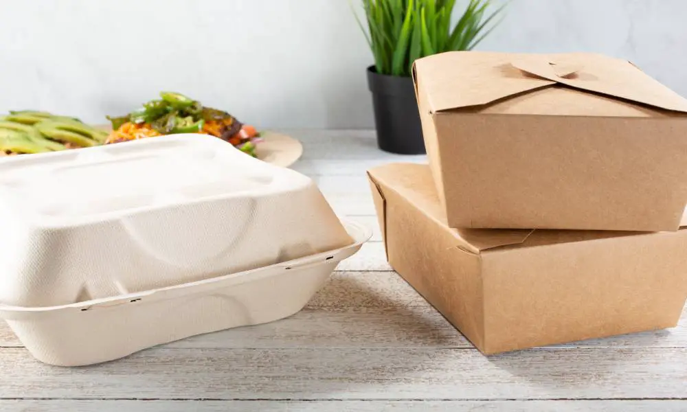 A set of paper to-go containers sitting on a table. There's a plate of food and a plant on the table behind the containers.