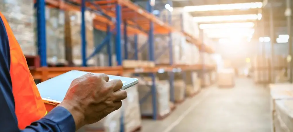A close-up view shows a person holding a silver tablet while standing in the aisle of a warehouse.
