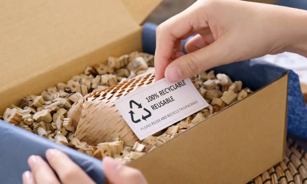 Two hands opening a cardboard box. One hand holds up a card in the box that reads “100% RECYCLABLE REUSABLE.”