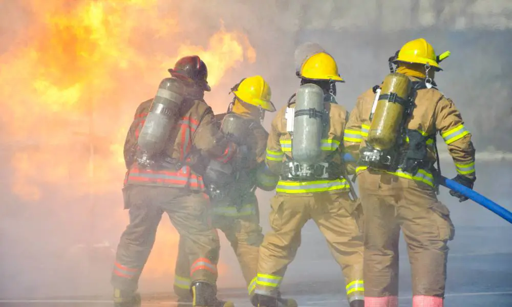 Four firefighters use a hose to put out a fire. One wears a red hard hat while the other wear yellow ones.