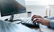 A man types on a keyboard. Next to him is another keyboard and monitor and a to-go coffee cup.