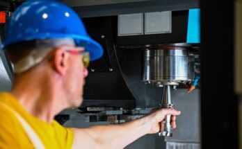 A man in a yellow shirt and blue hard hat reaching out with a tool to change the drill bit on a CNC machine.