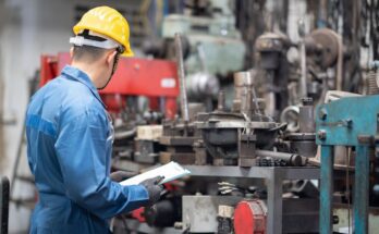 A worker wearing a blue work shirt and yellow hard hat holds a clipboard, looking over equipment at a factory.