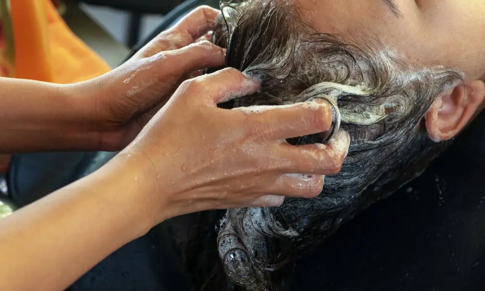 A salon employee washes a customer's hair over a shampoo bowl. Running water and suds fill the basin.