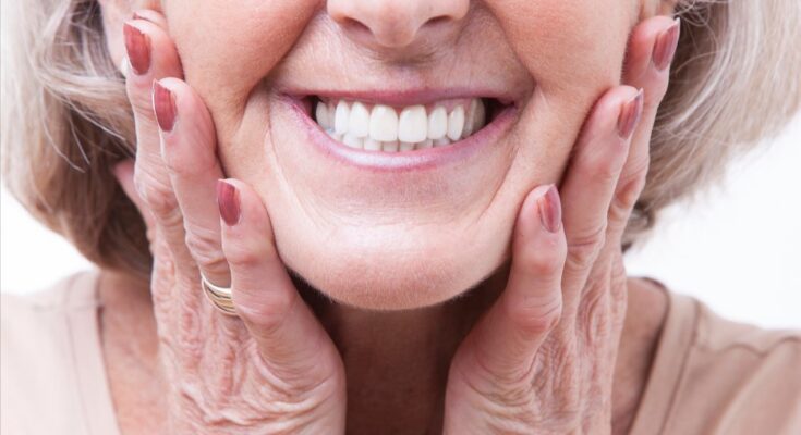 A close-up of an older woman with gray hair smiling widely and showing her teeth. Her hands are on the sides of her face.