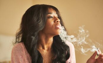 An African American woman sits and exhales smoke. She holds a cannabis joint and looks toward the right.