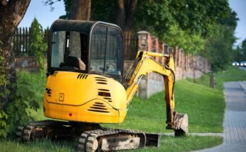 A black and yellow mini excavator sits on a patch of grass next to a tall, mature tree and brick fence.