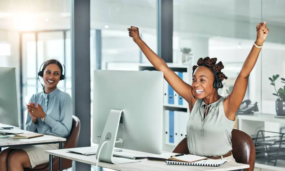 A woman sitting at her desk, raising her arms in the air and smiling. There's a woman at another desk smiling and clapping.