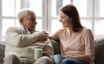 An old father and his young-adult daughter sit close together on a couch, holding mugs and smiling at each other.