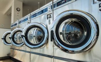 A row of numbered stainless steel commercial washing machines sit with their doors open in a laundromat.