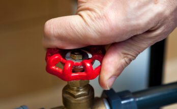 A close-up view shows a hand on the red knob of a brass water valve that links two blue water pipes.